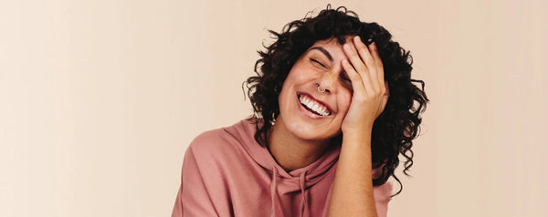 Person with curly hair and a nose piercing laughing while holding their face.